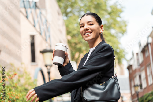 Trendy young woman with coffee mug walking through city streets in summer, great for themes like casual business style, weekend vibes, urban confidence, or influencer content