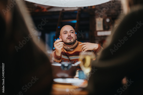 A man sits indoor at a bar wearing a warm sweater, actively talking with others. Surroundings are cozy with warm atmosphere. Focus on conversation and social interaction in a lively place.