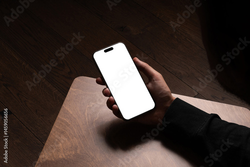 A person holding smartphone mockup on a wooden table in a dark cozy room with wooden floor. Isolated blank surface to place your design.