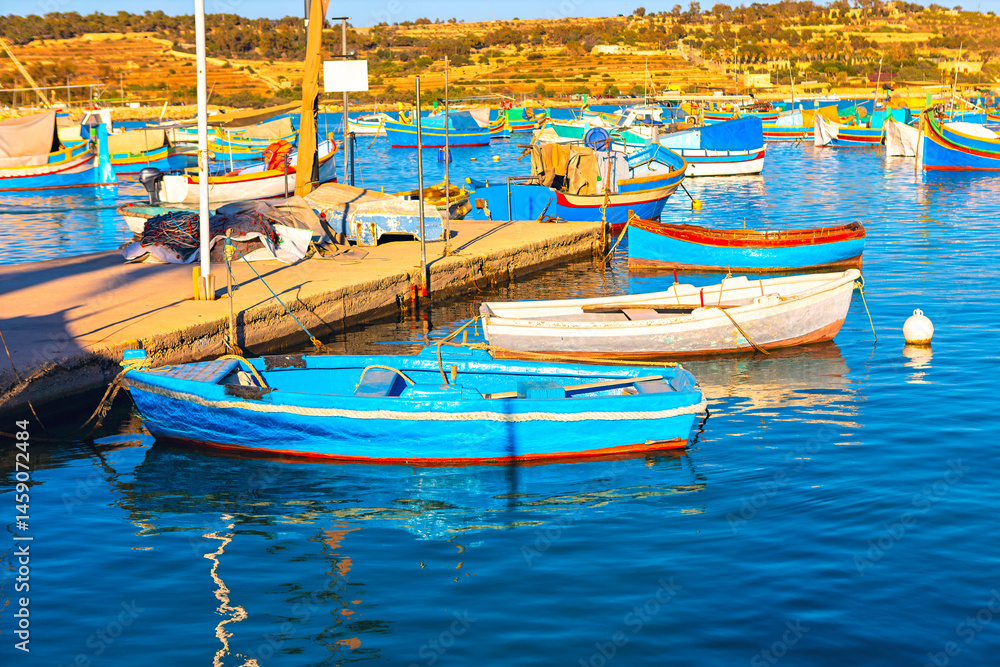 Serene harbor scene with numerous small boats, many painted in bright colors, docked along a wooden pier. In background, a hilly landscape with sparse vegetation stretches under a clear sky