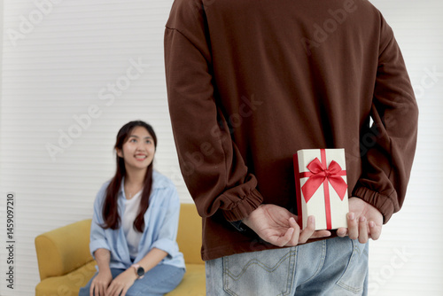 Man hiding gift box behind his back, ready to give anniversary present to lover girlfriend for celebration in living room. Happy Asian couple make surprise, love and happiness. Happy Valentine Day.