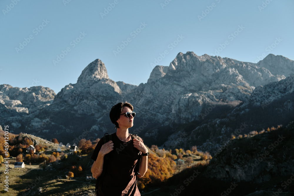Naklejka premium Female solo hiker with a backpack admires the majestic peaks of Montenegro in warm autumn light. The village below Bukumirsko Lake glows with vibrant fall colors in this cinematic mountain landscape.