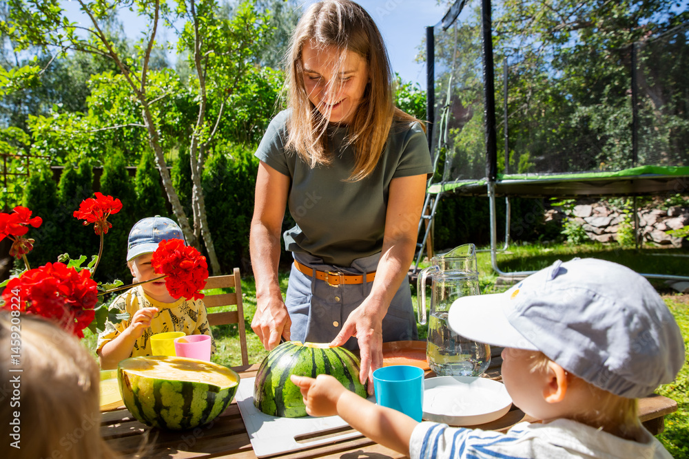Obraz premium Mother cutting watermelon with toddlers at a backyard table. Sunny summer day, family time, and cozy outdoor moment full of warmth.