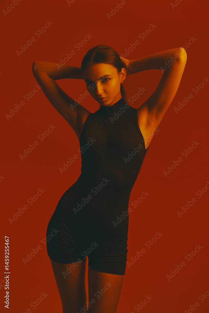 Portrait of attractive girl in black jumpsuit posing with confidence and elegance against red studio background in neon light. Concept of fashion, style, youth culture, trends