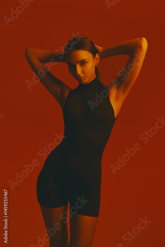 Portrait of attractive girl in black jumpsuit posing with confidence and elegance against red studio background in neon light. Concept of fashion, style, youth culture, trends