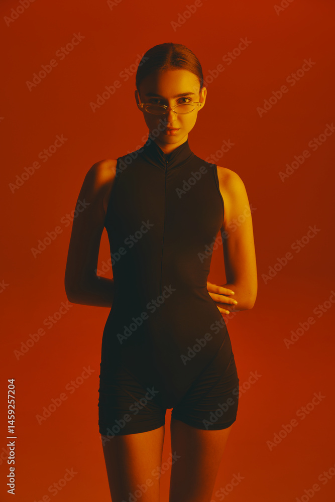 Portrait of confident and stylish young woman in black jumpsuit and trendy glasses, posing with intense gaze in camera against red background in neon. Concept of fashion, style, youth culture, trends