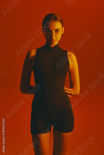 Obraz na płótnie Portrait of confident and stylish young woman in black jumpsuit and trendy glasses, posing with intense gaze in camera against red background in neon. Concept of fashion, style, youth culture, trends w salonie