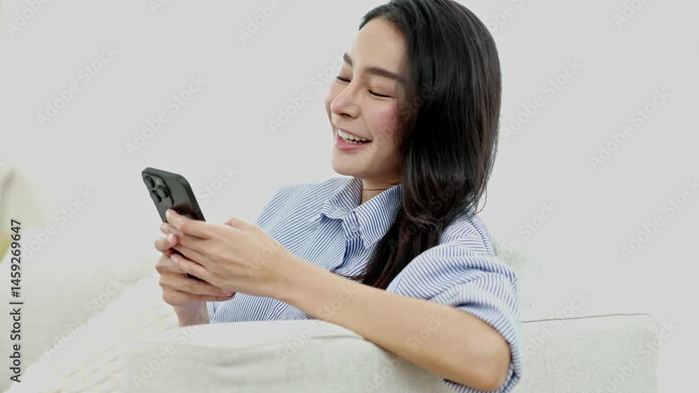 Asian Cheerful woman sitting on couch indoors, smiling and using smartphone in relaxed mood, enjoying technology and communication in cozy atmosphere. Asian Cheerful woman sitting on couch indoors, smiling and using smartphone in relaxed mood, enjoying technology and communication in cozy atmosphere.
