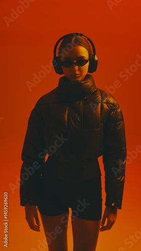 Obraz na płótnie Portrait of young woman wearing black shorts and winter jacket, sunglasses and headphones against red studio background in neon light. Concept of fashion, style, youth culture, trends w salonie