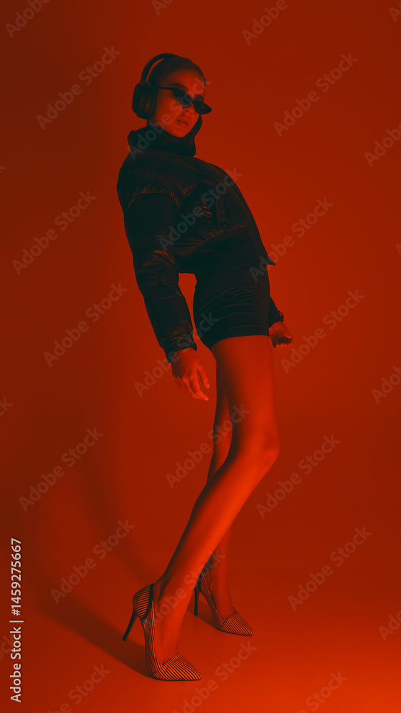 Stylish woman leans back, wearing heels, shorts, puffer jacket and sunglasses, in dynamic pose against red studio background in neon light. Concept of fashion, style, youth culture, trends