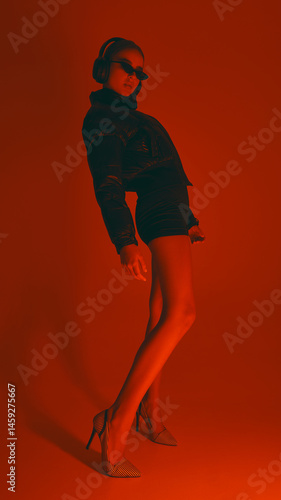Obraz na płótnie Stylish woman leans back, wearing heels, shorts, puffer jacket and sunglasses, in dynamic pose against red studio background in neon light. Concept of fashion, style, youth culture, trends w salonie