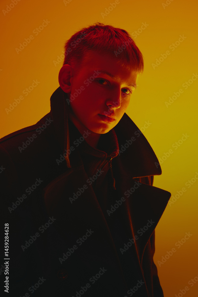 Portrait of young man wearing black coat, looking at camera, posing against yellow studio background in neon light. Concept of bold fashion, style, youth culture, self-expression
