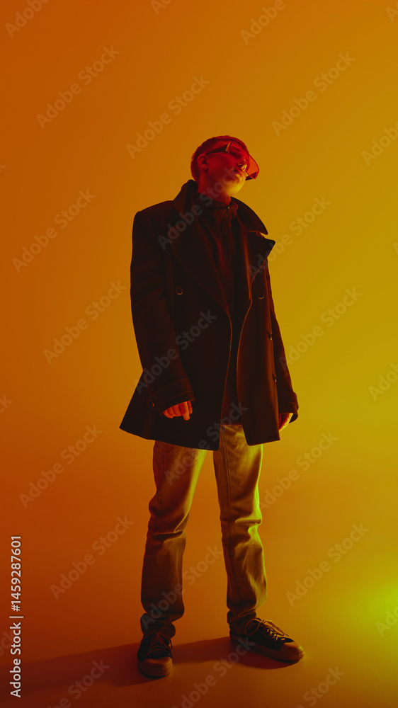 Full-length portrait of stylish, confident young man in jeans, black coat and trend red sunglasses, posing against yellow background in neon light. Concept of bold fashion, style, self-expression