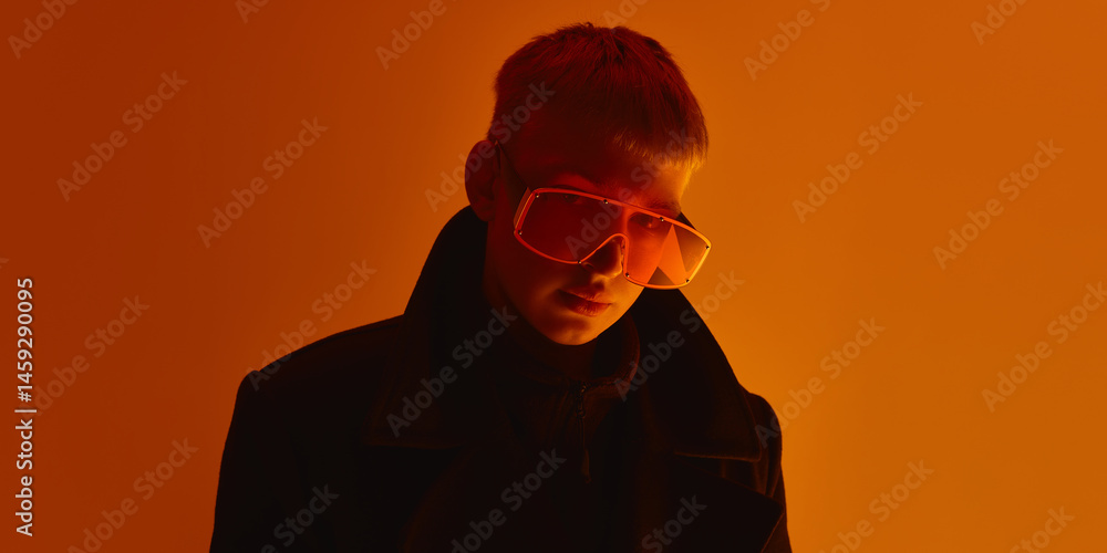 Close up of man wearing oversized orange sunglasses, looking seriously at camera under intense light against orange studio background. Concept of bold fashion, style, youth culture, self-expression