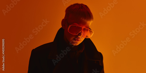 Close up of man wearing oversized orange sunglasses, looking seriously at camera under intense light against orange studio background. Concept of bold fashion, style, youth culture, self-expression