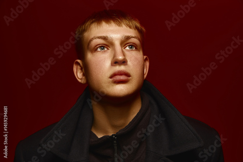 Close-up portrait of young man in dark jacket posing against rich red background, looking upwards with intense expression. Concept of contemporary fashion, youth culture, modern style