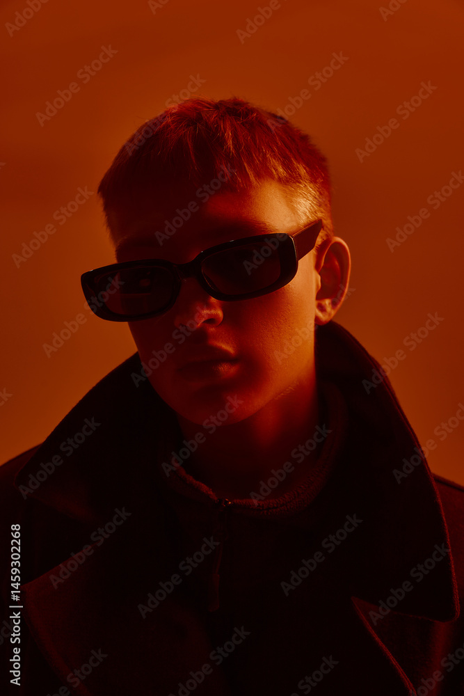 Close-up portrait of young stylish guy in trendy sunglasses and black coat, posing against red studio background in neon light. Concept of bold fashion, style, youth culture, self-expression