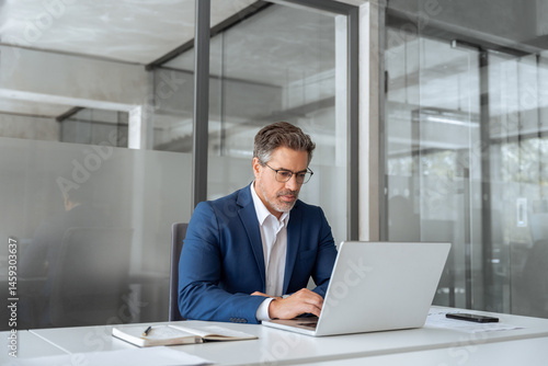 Mature indian ceo business man in suit using pc laptop for trading, looking focused at screen. Middle age latin male entrepreneur businessman working on financial work at desk in office. Copy space