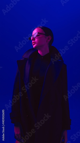 Obraz na płótnie Portrait of young confident woman wearing sunglasses and black coat, posing against blue background in neon light. Concept of bold fashion, style, youth culture, self-expression w salonie