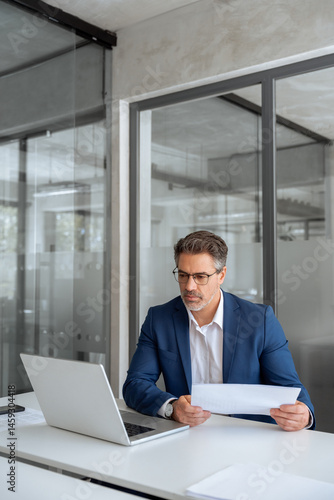 Entrepreneur manager businessman banking analyst doing paperwork analysing using pc. Busy latin mature business man working on laptop computer reading financial document report in office. Vertical 
