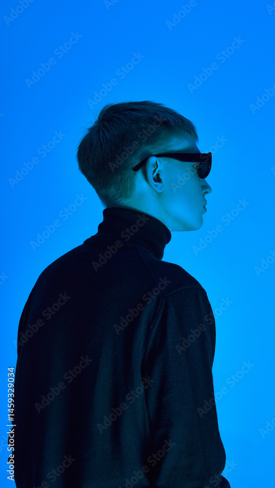 Side profile of stylish young man in turtleneck and sunglasses, posing under deep neon blue background ,expresses futuristic elegance. Concept of bold fashion, style, youth culture, self-expression