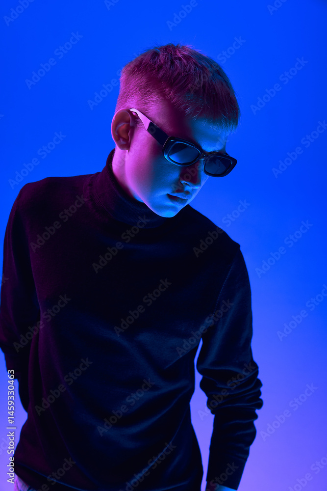 Portrait of stylish young guy in sunglasses and black turtleneck posing against blue background in neon light. Concept of bold fashion, style, youth culture, self-expression