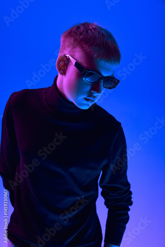 Obraz na płótnie Portrait of stylish young guy in sunglasses and black turtleneck posing against blue background in neon light. Concept of bold fashion, style, youth culture, self-expression w salonie