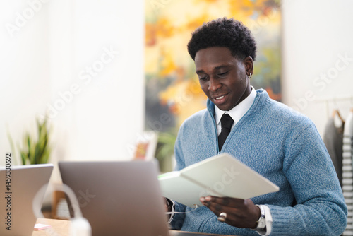 A young professional man reviews notes in a book at his desk. He is wearing a blue sweater and tie, working from his home office.