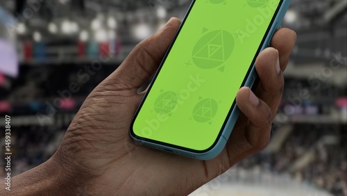 GREEN SCREEN CHROMA KEY A hand holding a phone with a green screen at a hockey game in an indoor stadium, ideal for showcasing live sports or betting apps
