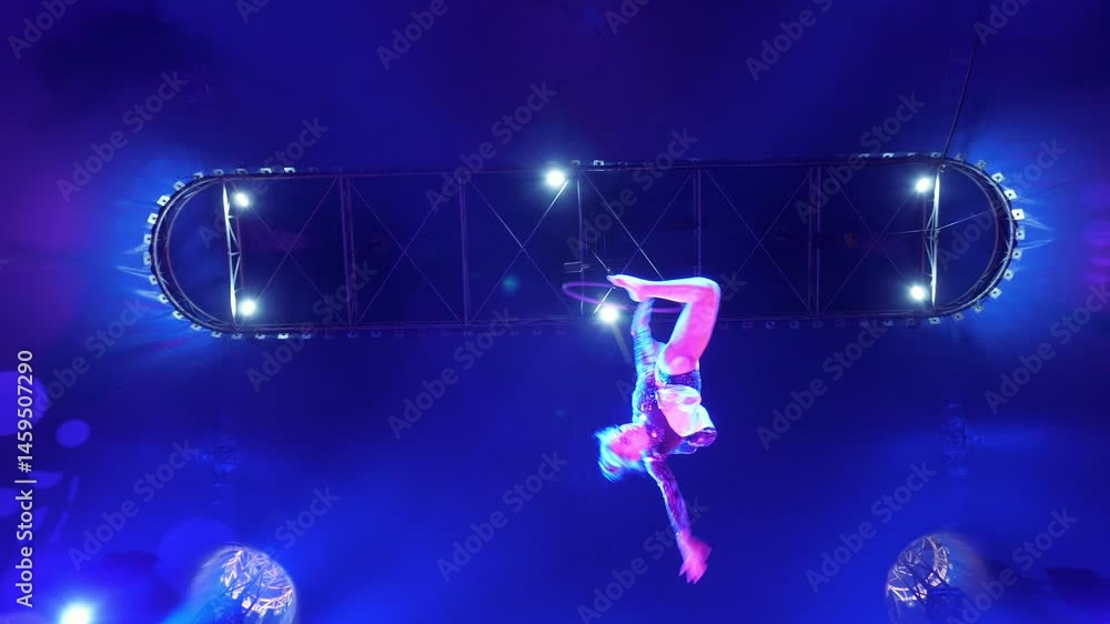A flexible aerial gymnast quickly rotates around herself on a gymnastic hoop under the dome of the circus. Performance of an aerial gymnast under the light of blue spotlights. A circus show. A flexible aerial gymnast quickly rotates around herself on a gymnastic hoop under the dome of the circus. Performance of an aerial gymnast under the light of blue spotlights. A circus show.