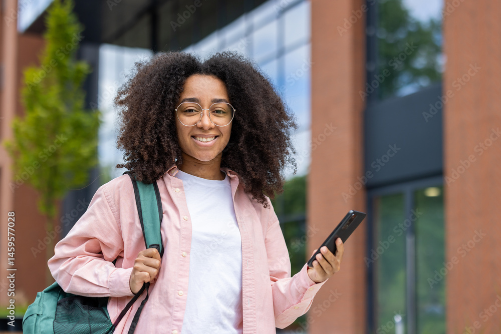 A smiling young woman stands outside, holding a phone and a backpack. She is wearing glasses and a pink shirt.