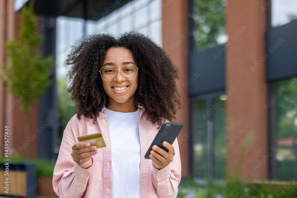 a smiling young woman holding a credit card and smartphone outdoors near a building with glass windows and modern architecture.