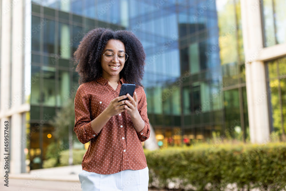 A smiling african american woman wearing glasses checks her smartphone in an urban setting near a business building.
