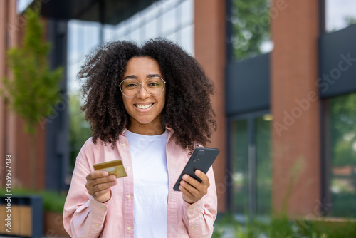 Obraz na płótnie a smiling young woman holding a credit card and smartphone outdoors near a building with glass windows and modern architecture. w salonie