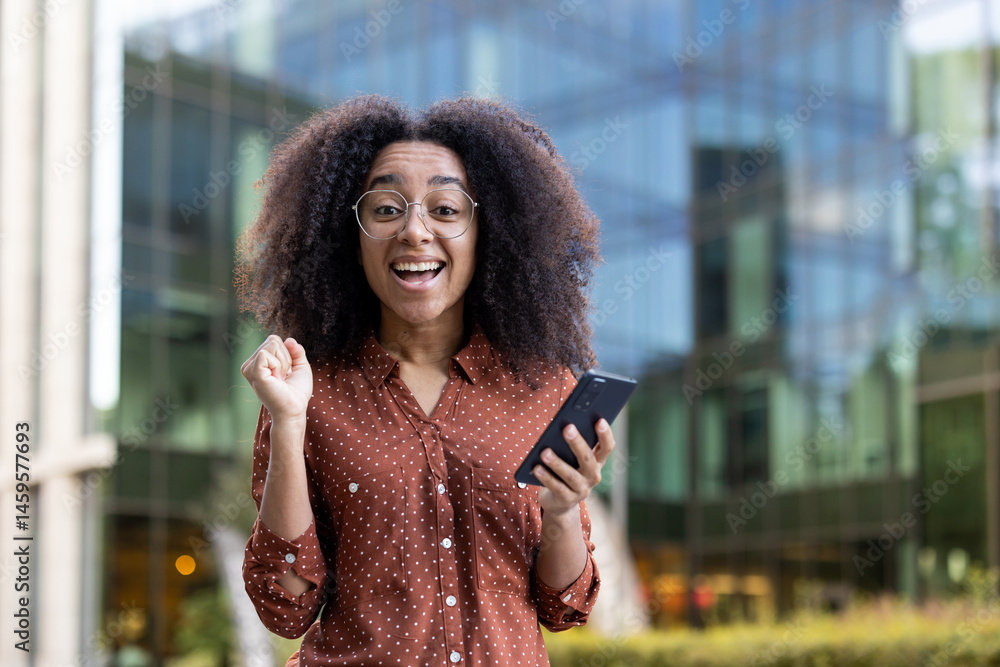 Excited young woman looks at her phone and celebrates a good news. A happy lady with glasses outside in front of a modern building.