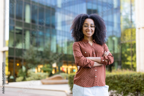 Obraz na płótnie A smiling black woman stands confidently in front of a modern building, arms crossed, ready for the day. w salonie