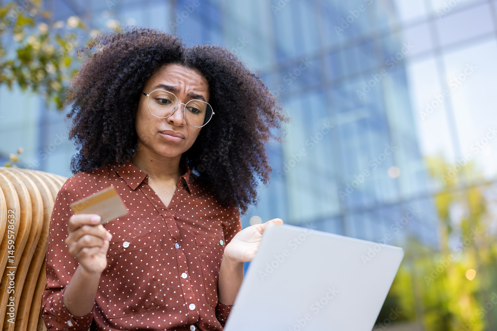 A young woman with curly hair looks puzzled while holding a credit card and using a laptop outdoors near a modern building.