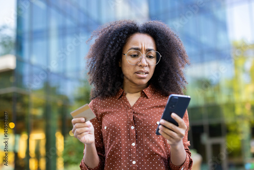 Obraz na płótnie A concerned woman in eyeglasses looks at her phone and credit card, possibly experiencing a payment issue outdoors. w salonie