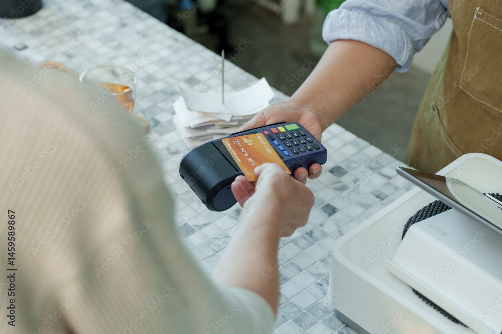 Customer tapping orange credit card on payment terminal at cafe counter demonstrating contactless payment and cashless transaction in modern small coffee shop Customer tapping orange credit card on payment terminal at cafe counter demonstrating contactless payment and cashless transaction in modern small coffee shop