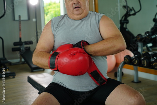 Elderly Caucasian man with a fat belly wearing gym clothes wearing red gloves practicing boxing and punching doing exercises for health and lose weight in a fitness centere.