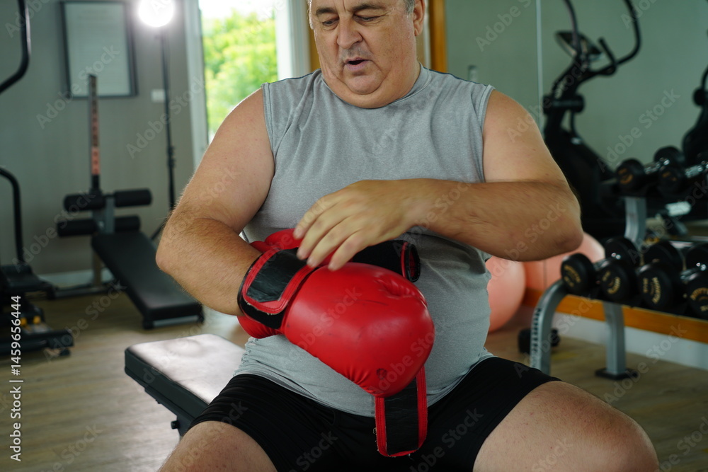 Elderly Caucasian man with a fat belly wearing gym clothes wearing red gloves practicing boxing and punching doing exercises for health and lose weight in a fitness centere.