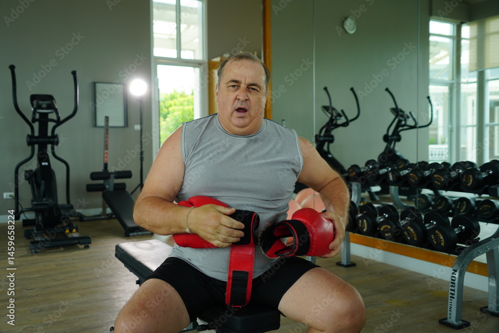 Elderly Caucasian man with a fat belly wearing gym clothes wearing red gloves practicing boxing and punching doing exercises for health and lose weight in a fitness centere.