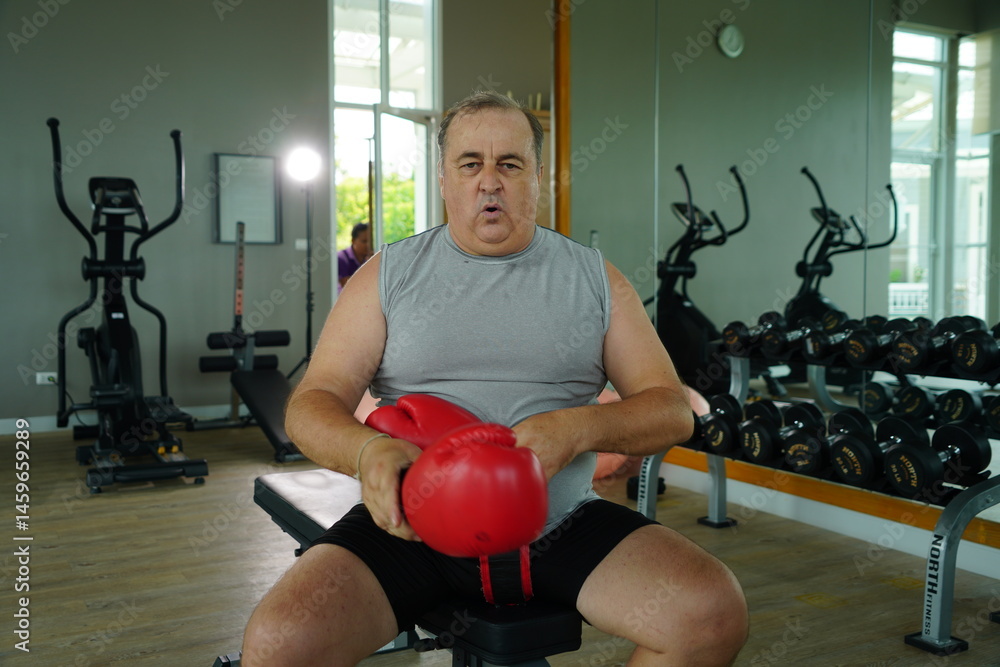 Elderly Caucasian man with a fat belly wearing gym clothes wearing red gloves practicing boxing and punching doing exercises for health and lose weight in a fitness centere.