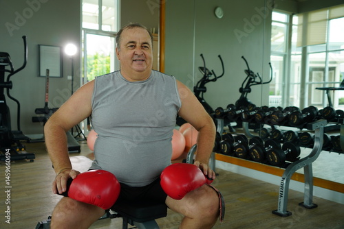 Elderly Caucasian man with a fat belly wearing gym clothes wearing red gloves practicing boxing and punching doing exercises for health and lose weight in a fitness centere.