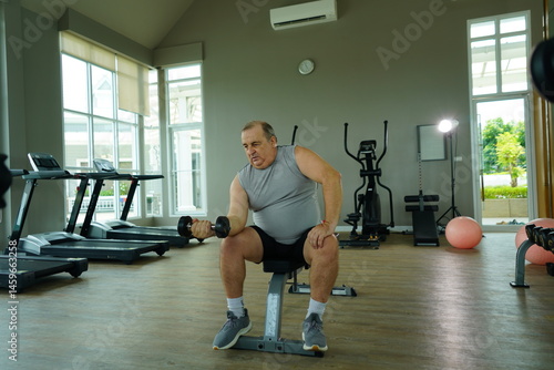 Elderly Caucasian man with a fat belly wearing gym clothes lifting dumbbells doing exercises to build muscle for health and lose weight in a fitness center Health and wellness concept