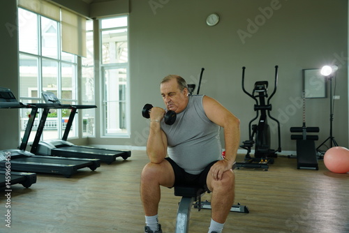 Obraz na płótnie Elderly Caucasian man with a fat belly wearing gym clothes lifting dumbbells doing exercises to build muscle for health and lose weight in a fitness center Health and wellness concept w salonie