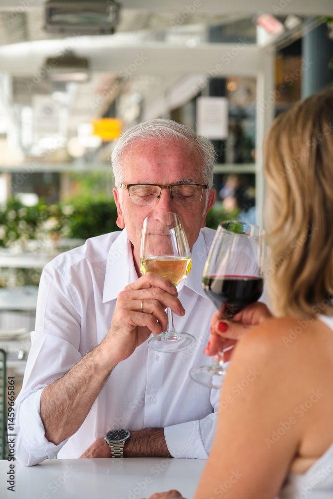 Senior man smelling a glass of white wine with closed eyes, a woman is drinking red wine next to him