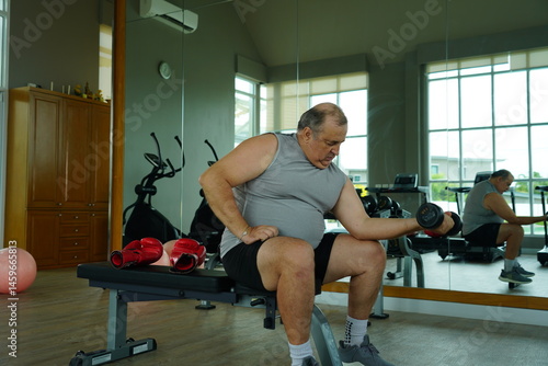 Obraz na płótnie Elderly Caucasian man with a fat belly wearing gym clothes lifting dumbbells doing exercises to build muscle for health and lose weight in a fitness center Health and wellness concept w salonie