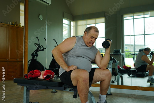 Obraz na płótnie Elderly Caucasian man with a fat belly wearing gym clothes lifting dumbbells doing exercises to build muscle for health and lose weight in a fitness center Health and wellness concept w salonie