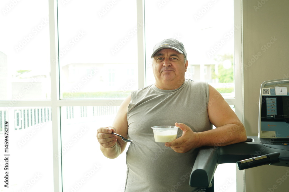 Fat Caucasian senior man with potbelly after workout hungry eating breakfast yoghurt,fruit salad healthy food and weight loss in fitness club. Health and wellness concept.